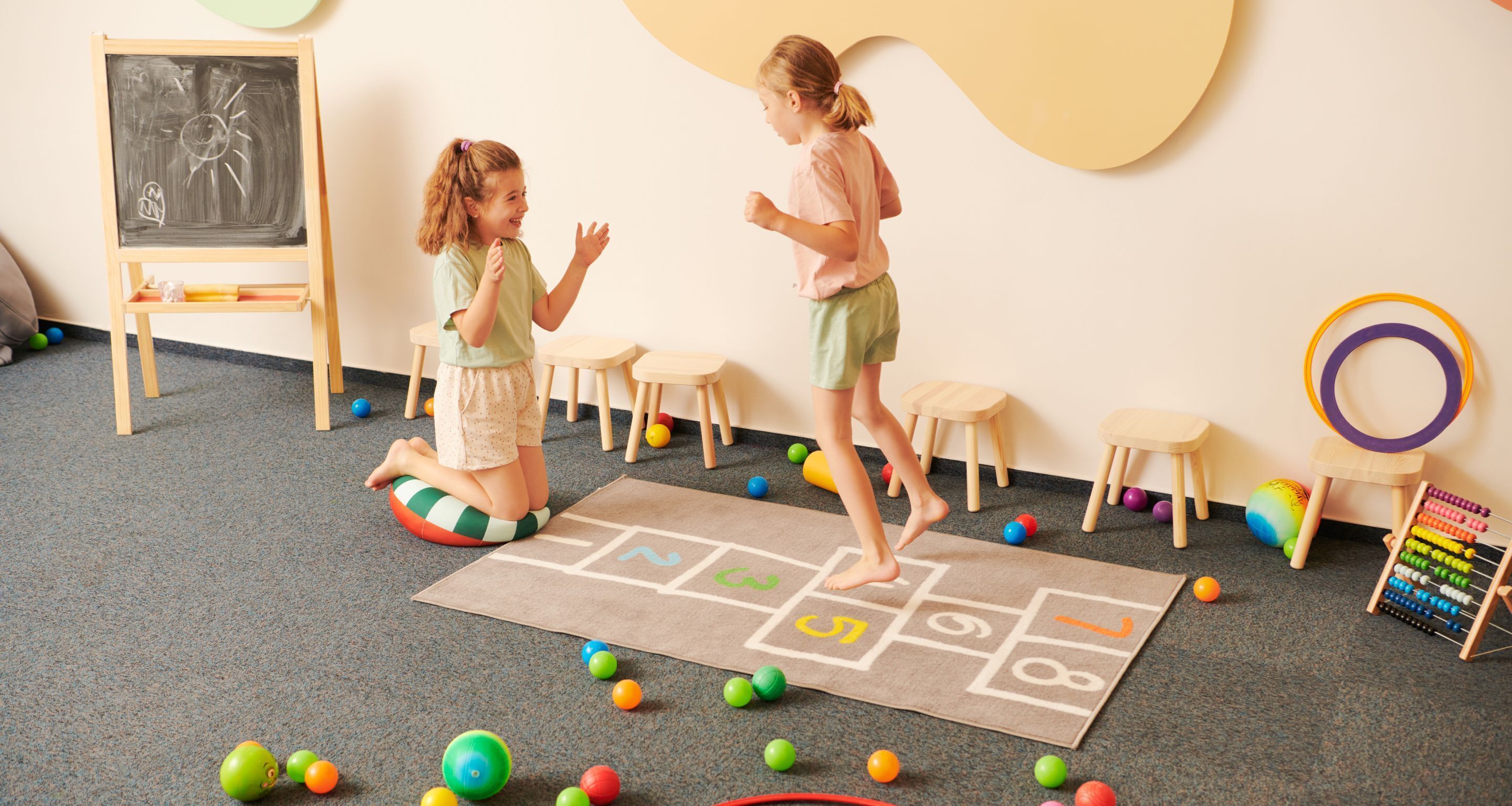 Two children playing hopscotch in a room with a chalkboard, wooden stools, and an abacus.