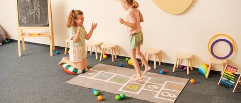 Two children playing hopscotch in a room with a chalkboard, wooden stools, and an abacus.