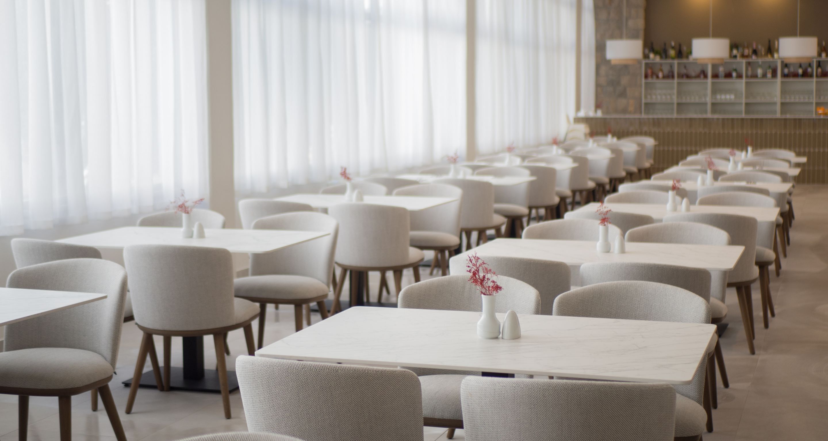 Un restaurant avec des rangées de tables et de chaises, de grandes fenêtres ornées de rideaux légers et un bar situé au fond de la salle.
