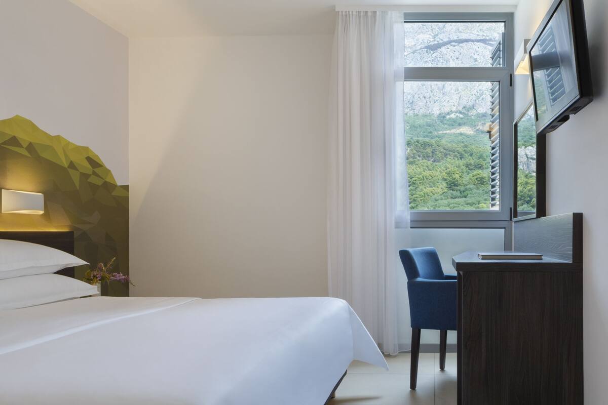 A hotel room with a bed, a desk, a chair, a TV, and a window with a view of greenery.
