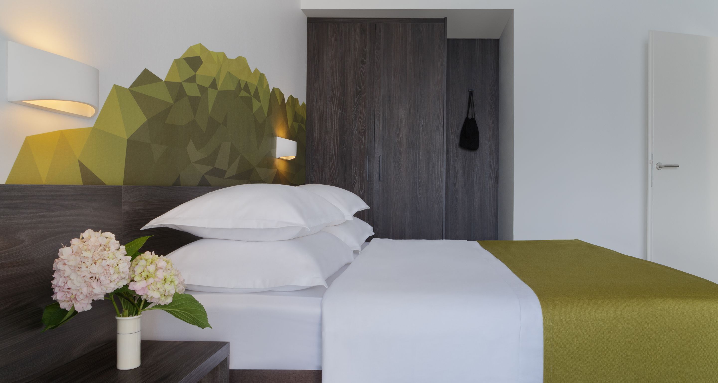 A hotel room with a bed, pillows, a bedside table with a vase of flowers, a wall-mounted lamp, and a geometric wall design.