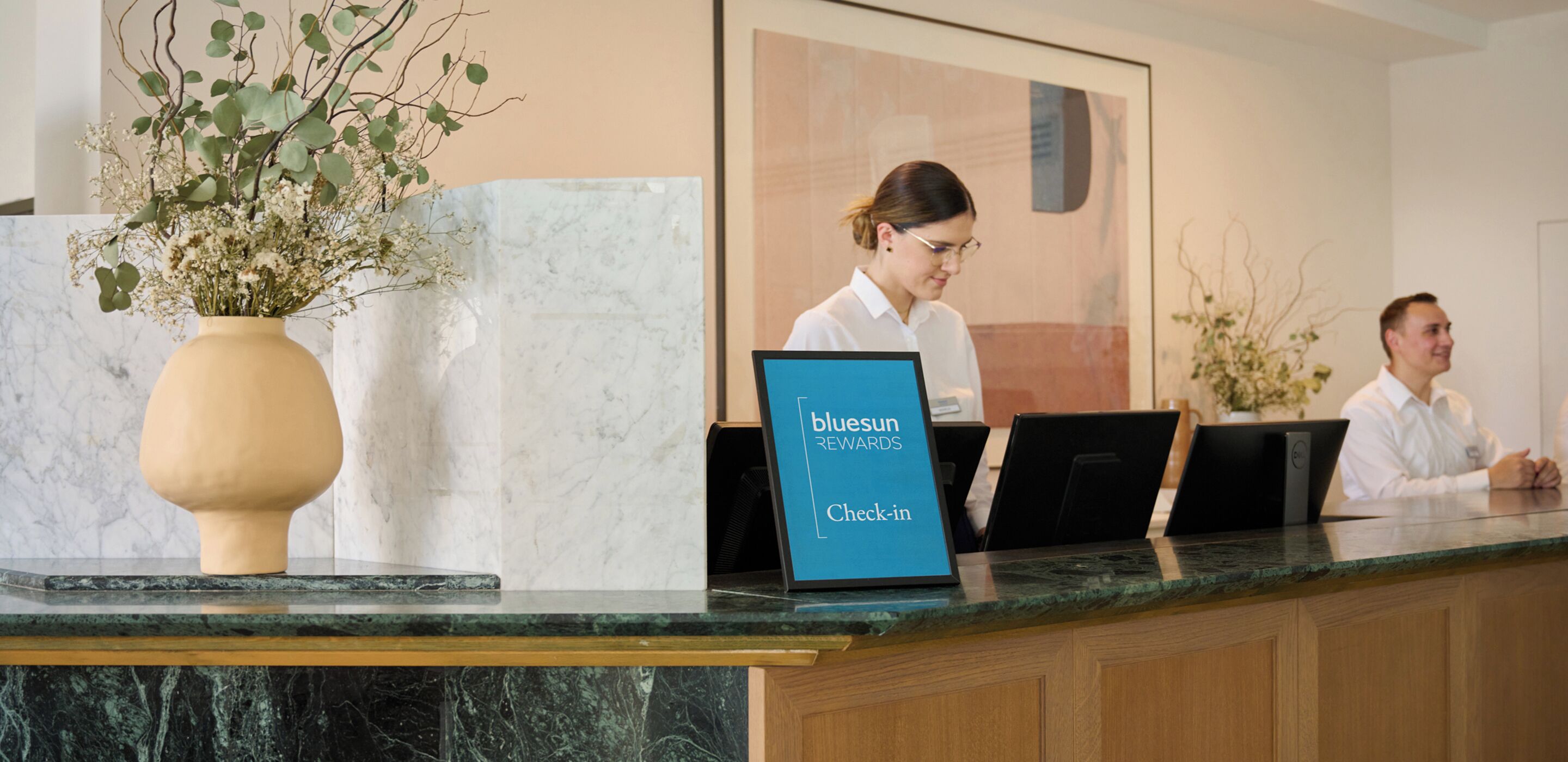 A hotel reception desk with a marble counter, a vase with flowers, and a sign for 'bluesun rewards check-in'.
