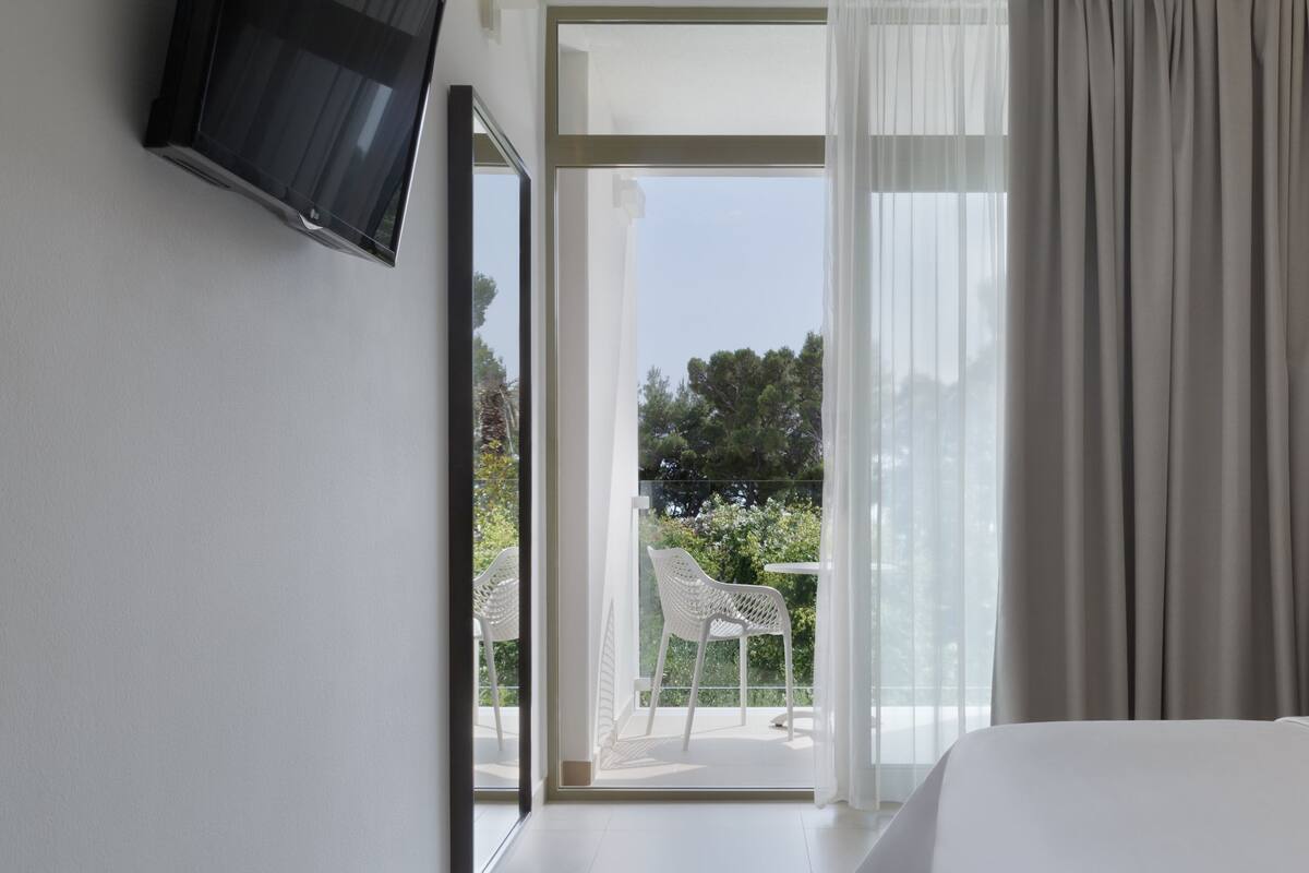 A hotel room with a bed, a TV mounted on the wall, a balcony with a chair, and a view of greenery outside.