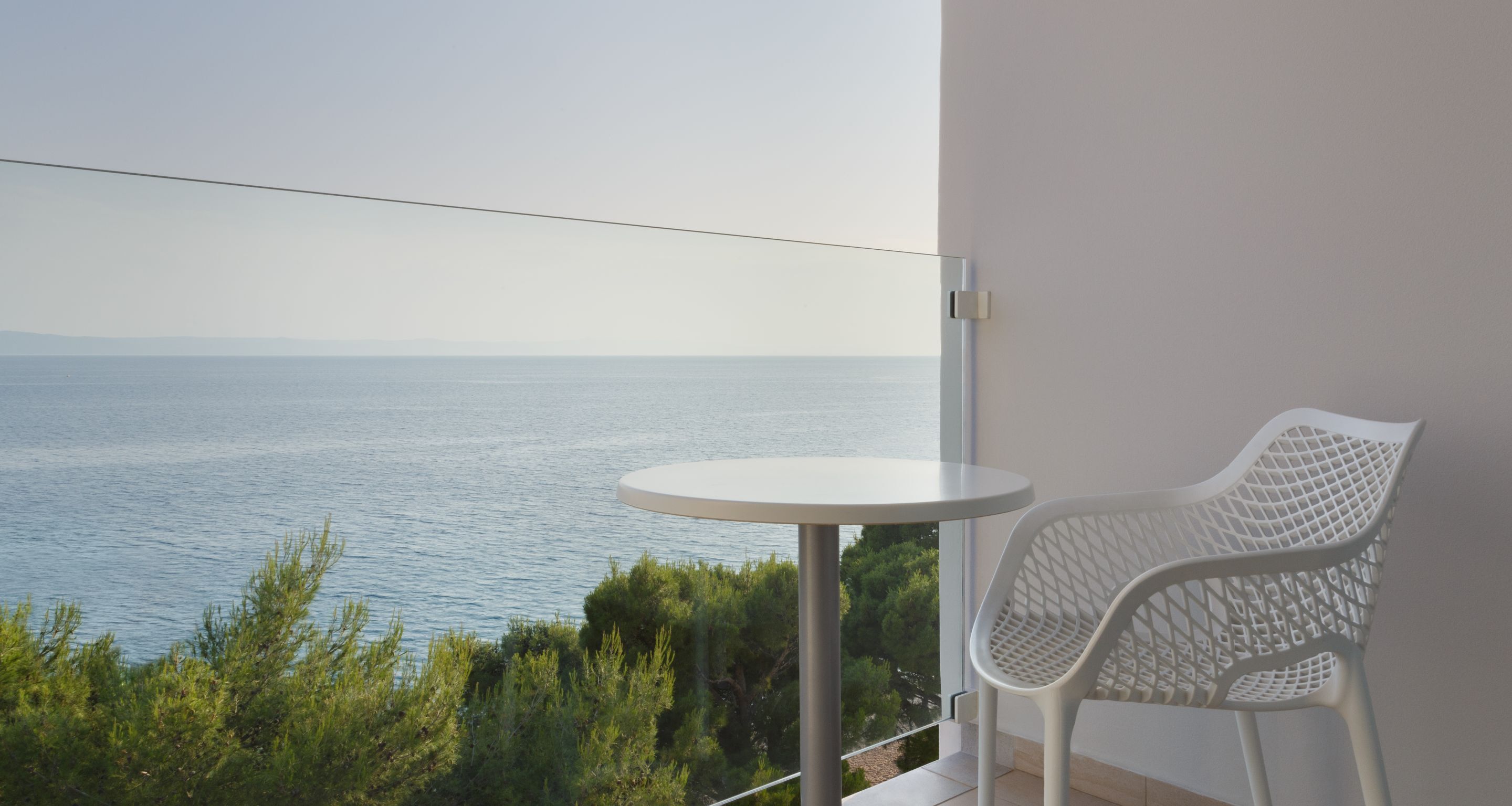 Un balcone con tavolo e sedia bianchi, affaccia su uno specchio d'acqua e vegetazione.
