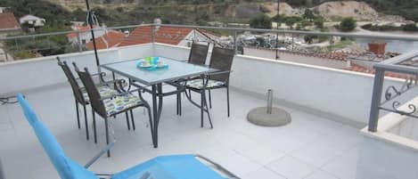 A terrace with a table and chairs, a blue umbrella, and a view of a hilly landscape.