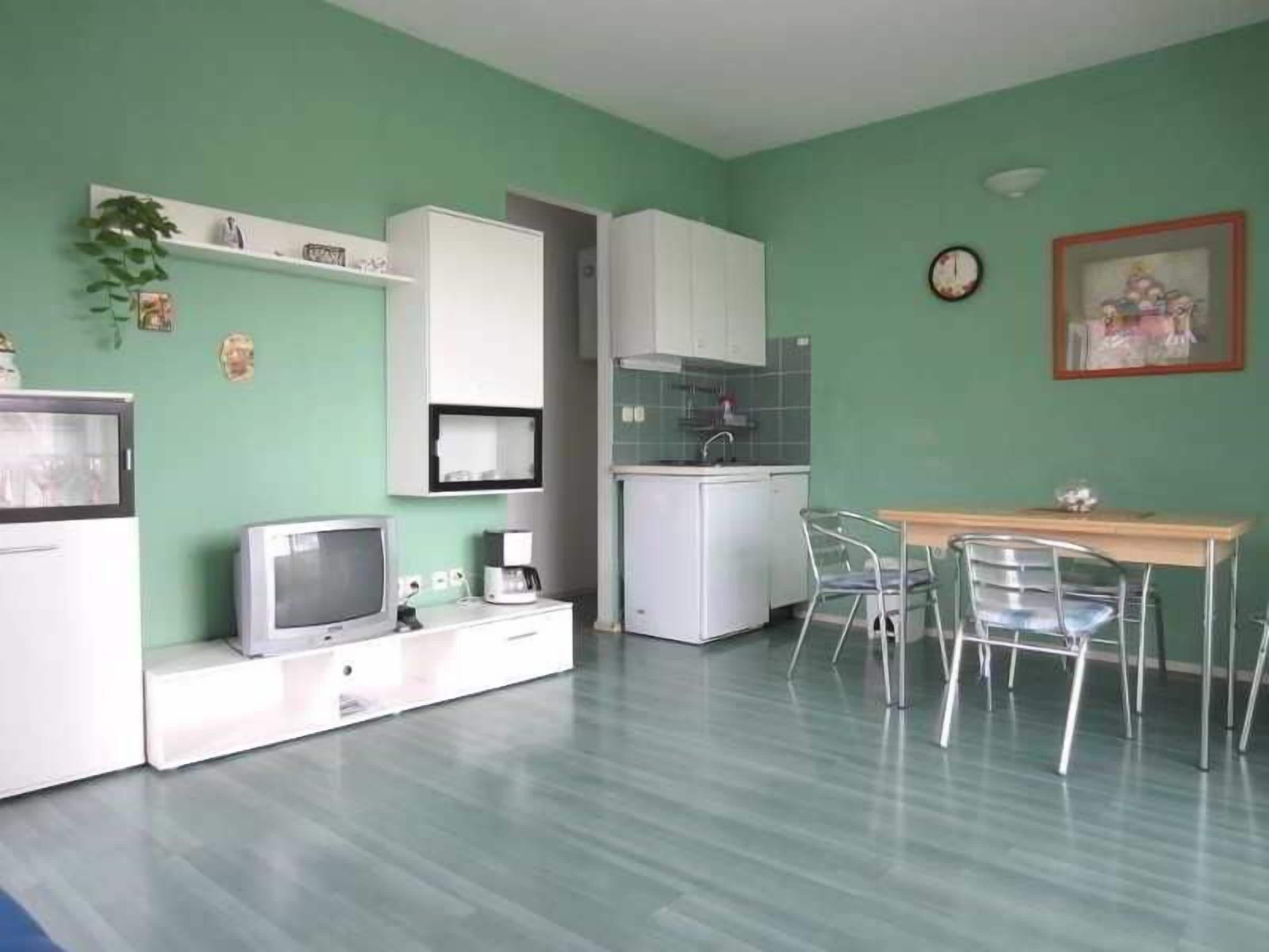 A compact kitchen with white cabinets, a dining table with chairs, a small refrigerator, and a television.