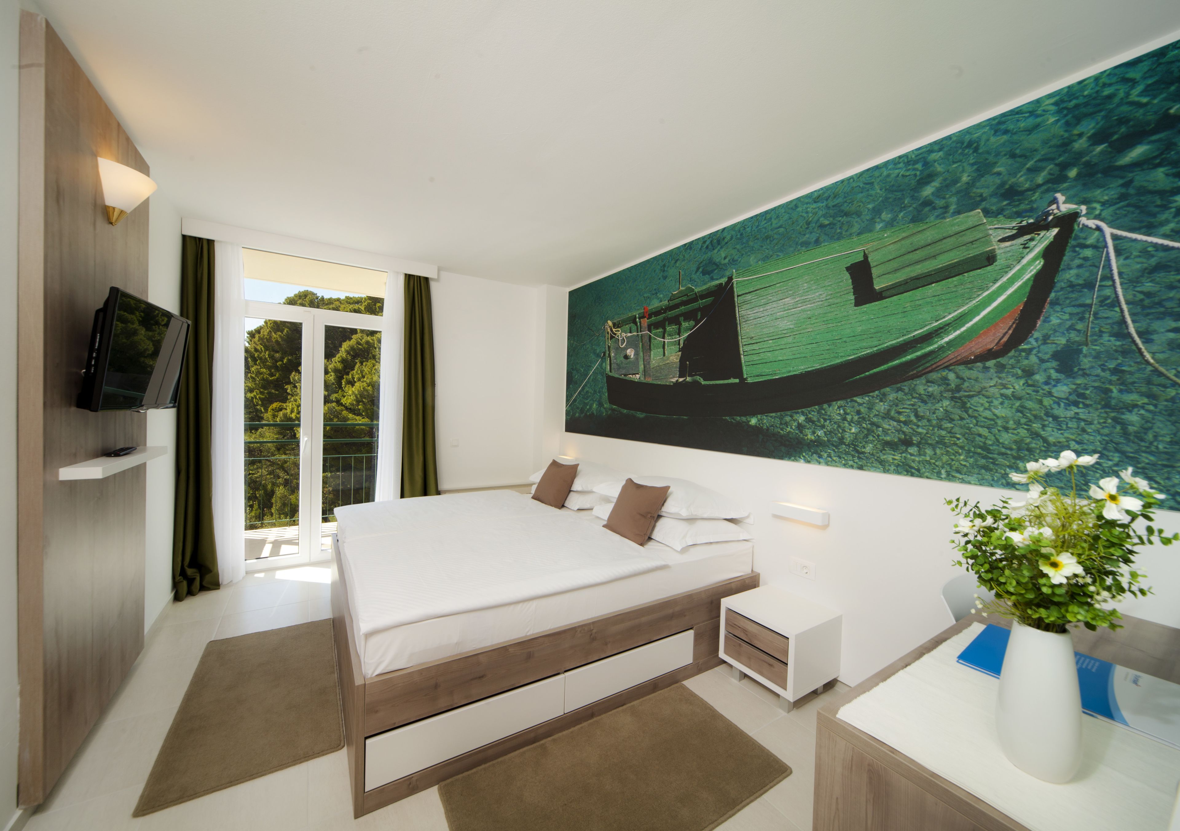 Une chambre d’hôtel moderne avec un grand lit, un téléviseur à écran plat, un tableau représentant un bateau et un bureau agrémenté d’un vase de fleurs.