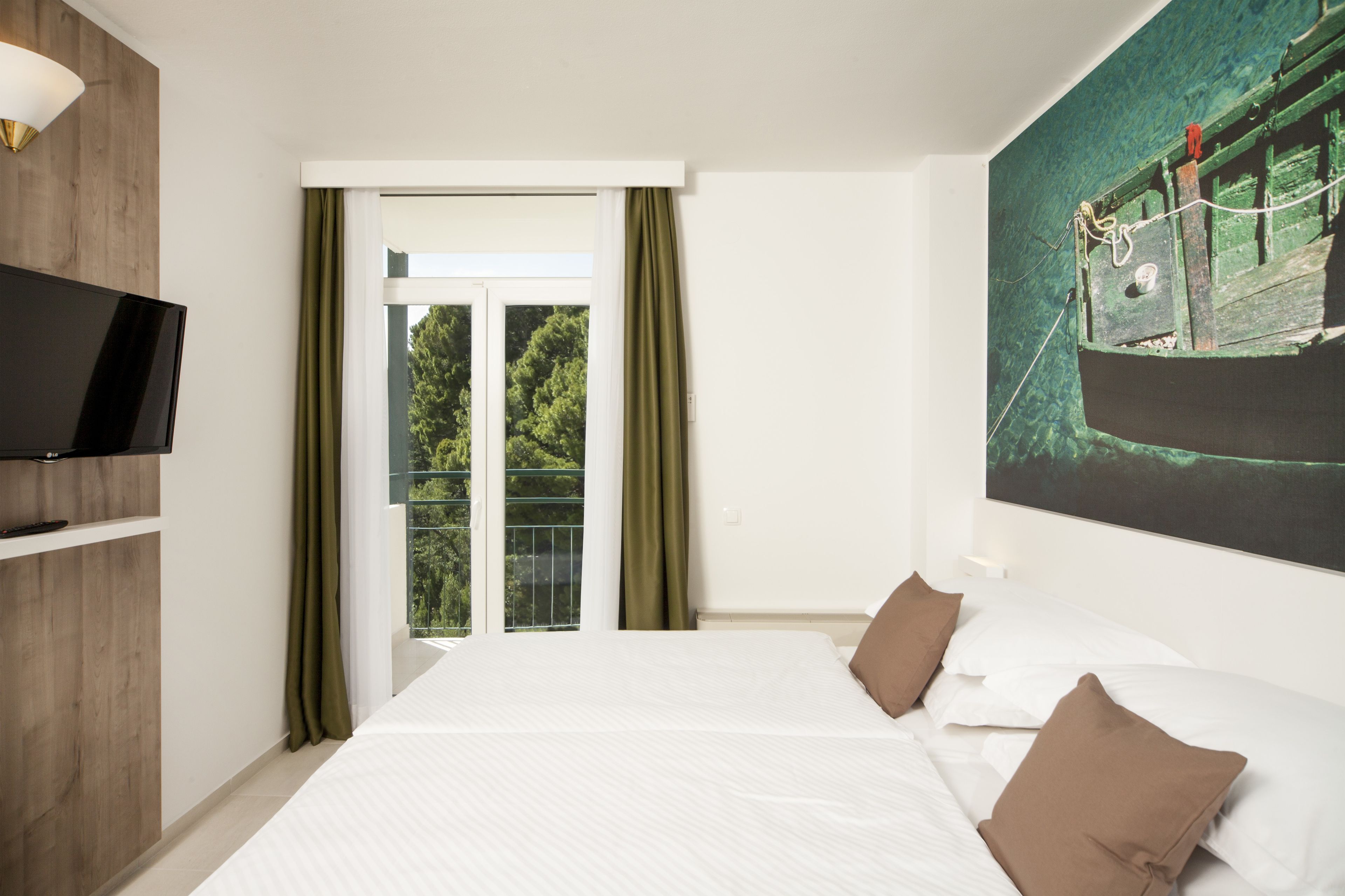 Ein Hotelzimmer mit einem großen Bett, einem Fernseher links und einem Balkon mit Blick auf Grünflächen.