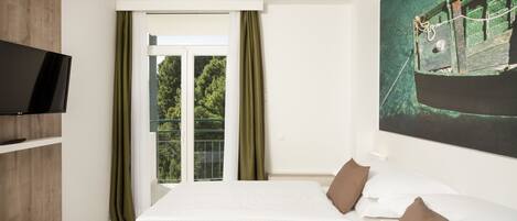A hotel room with a large bed, a television on the left, and a balcony with a view of greenery.