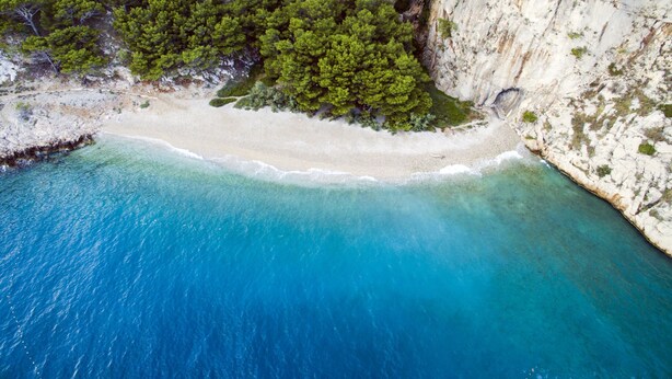 An aerial view of a coastal area with a sandy beach, clear blue waters, and dense green vegetation on the cliffs. - Bluesun Hotel Neptun (Tucepi)