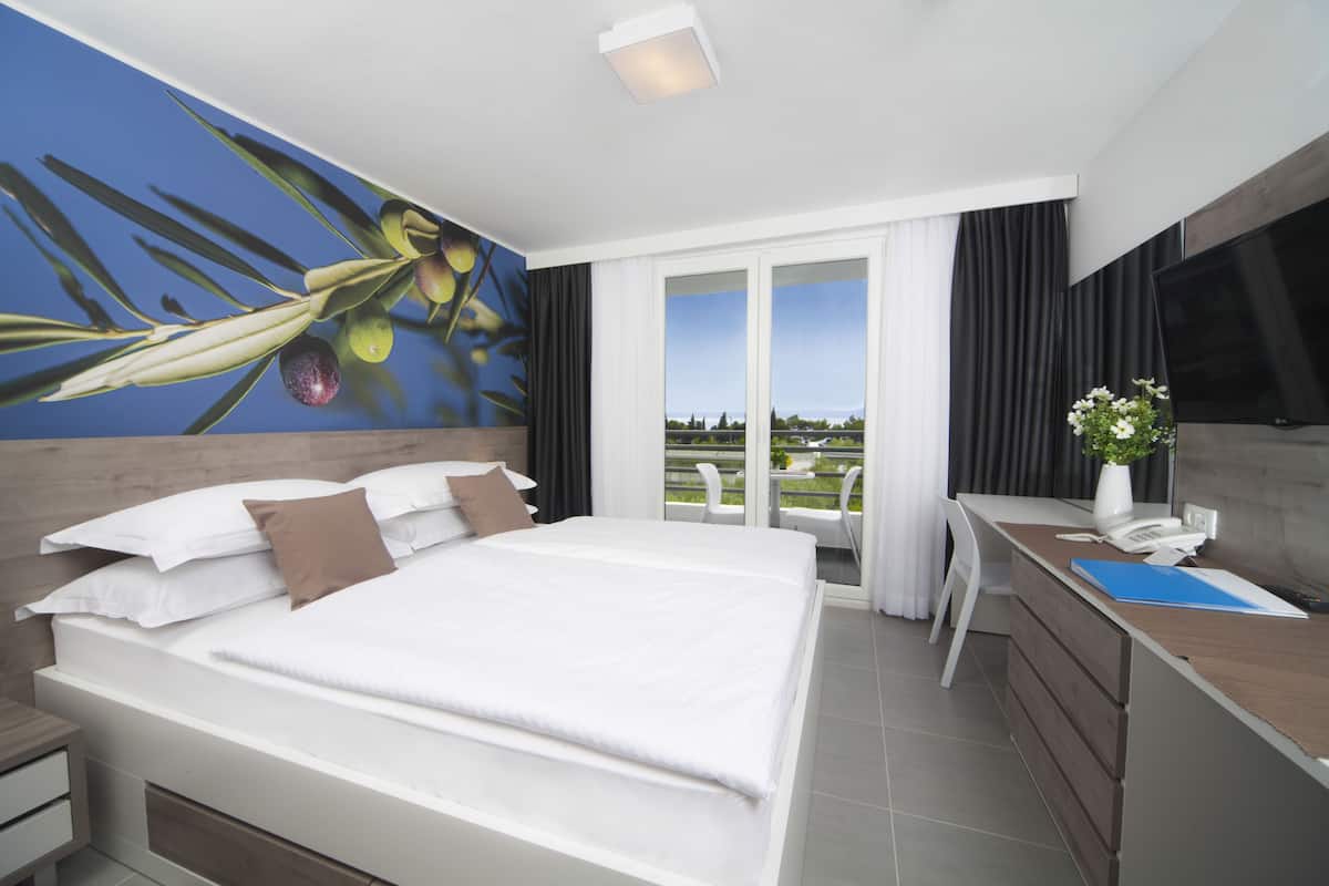 A modern hotel room with a large bed, a desk with a computer, a flat-screen TV, and a balcony with a view.
