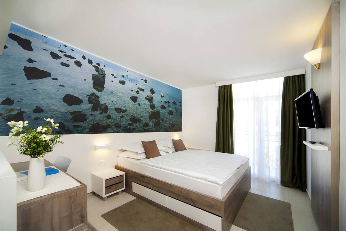 A modern hotel room with a large bed, a desk with a vase of flowers, a flat-screen TV, and a painting of a rocky seascape on the wall.