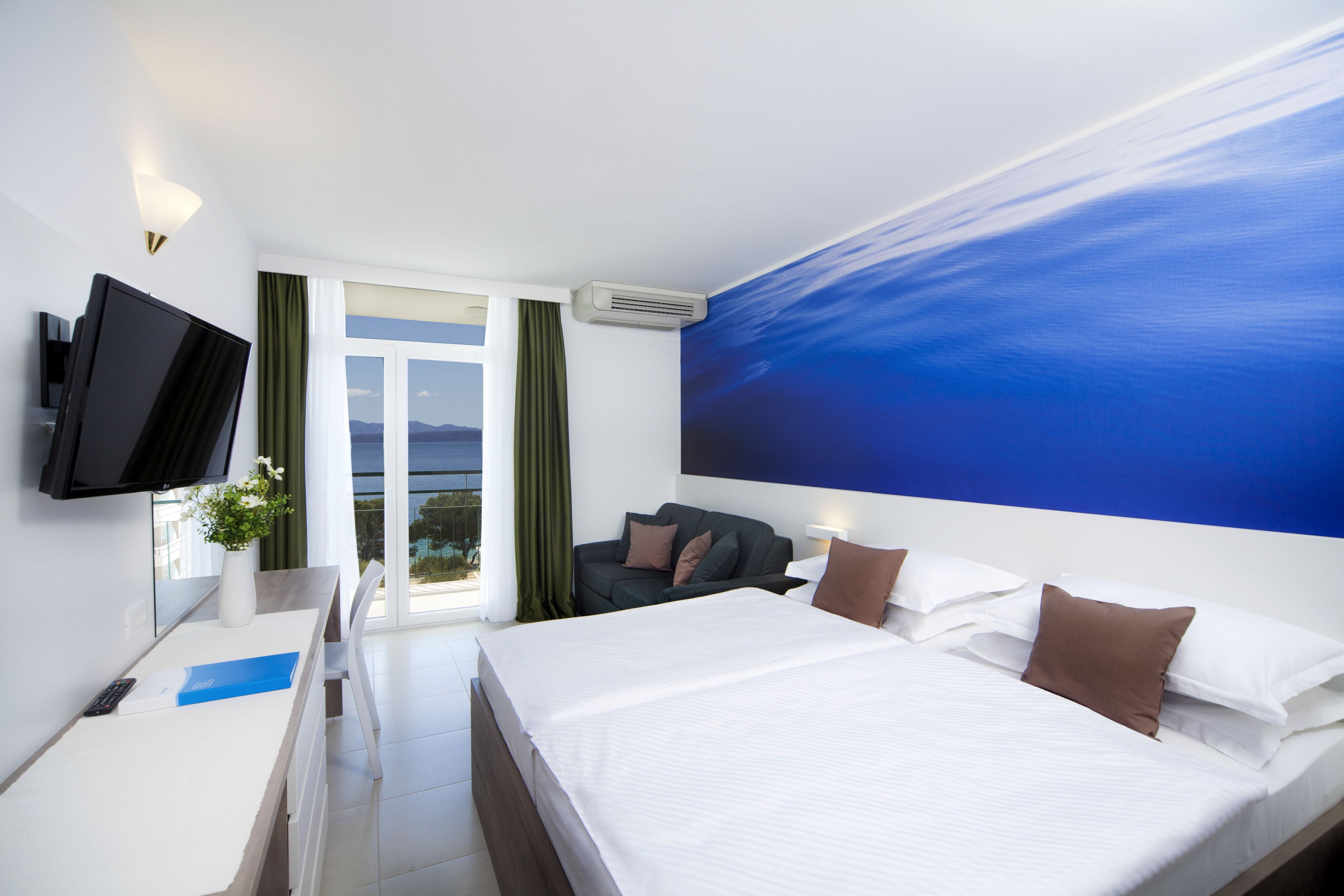 A hotel room with a bed, a desk, a TV, a window with curtains, and a wall with a blue mural.