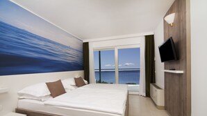 A hotel room with a large bed, a TV mounted on the wall, and a view of the sea through the window. - Bluesun Hotel Neptun (Tucepi)