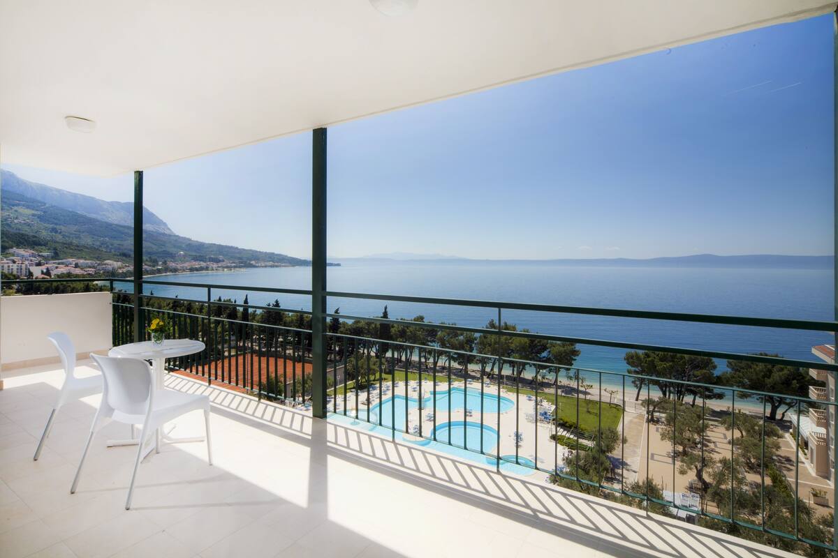 A balcony with a view of the sea, a swimming pool, and a table with chairs.