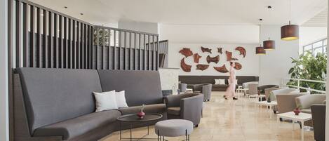 Een moderne lobby met een grijze hoekbank, een ronde salontafel en een pouf. Er staan meerdere fauteuils en een trap met een leuning.
