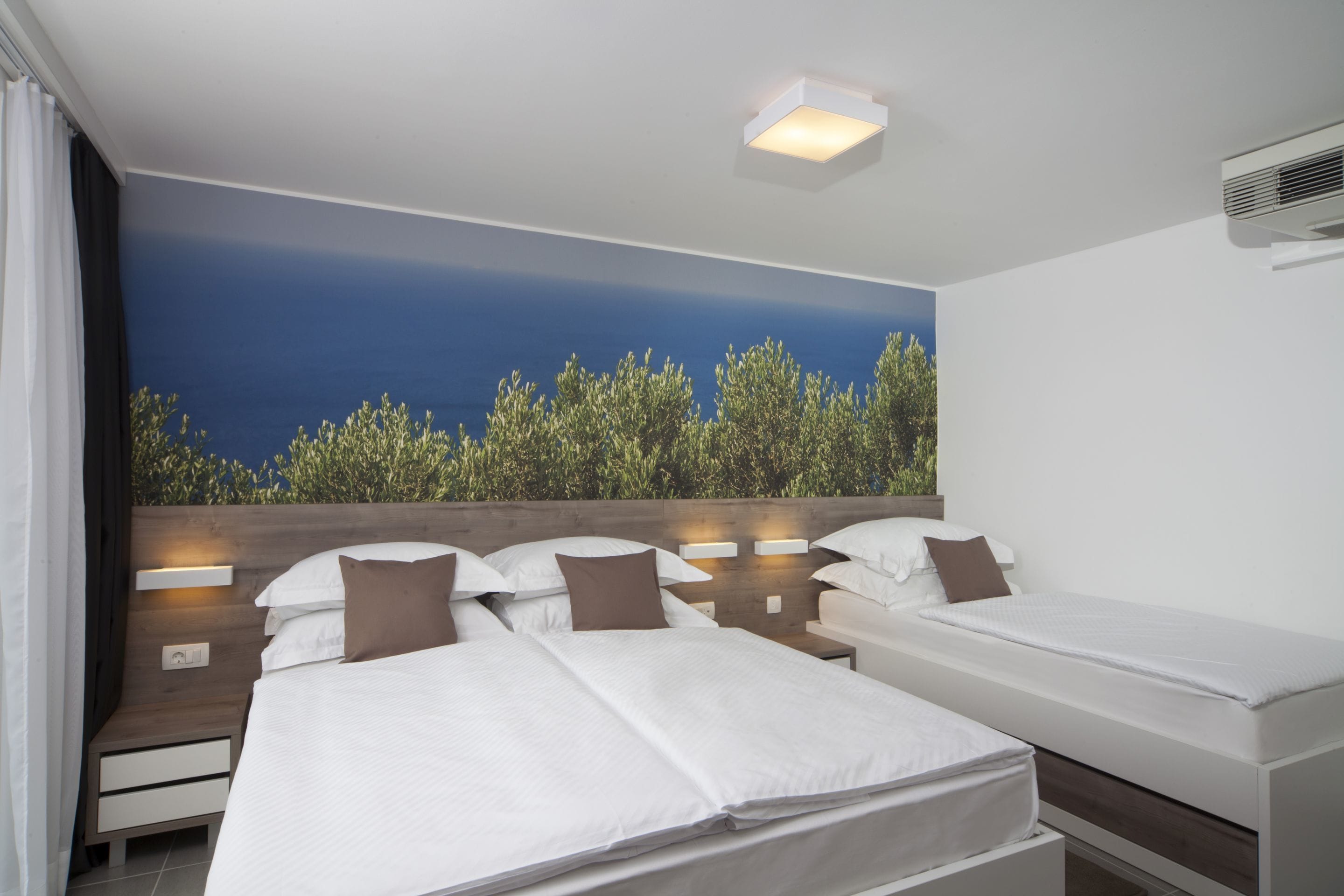 Una habitación de hotel con dos camas, un ventanal con vistas a árboles y una lámpara en el techo.