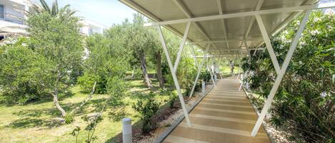 A covered walkway with a geometric metal framework, surrounded by greenery and trees.