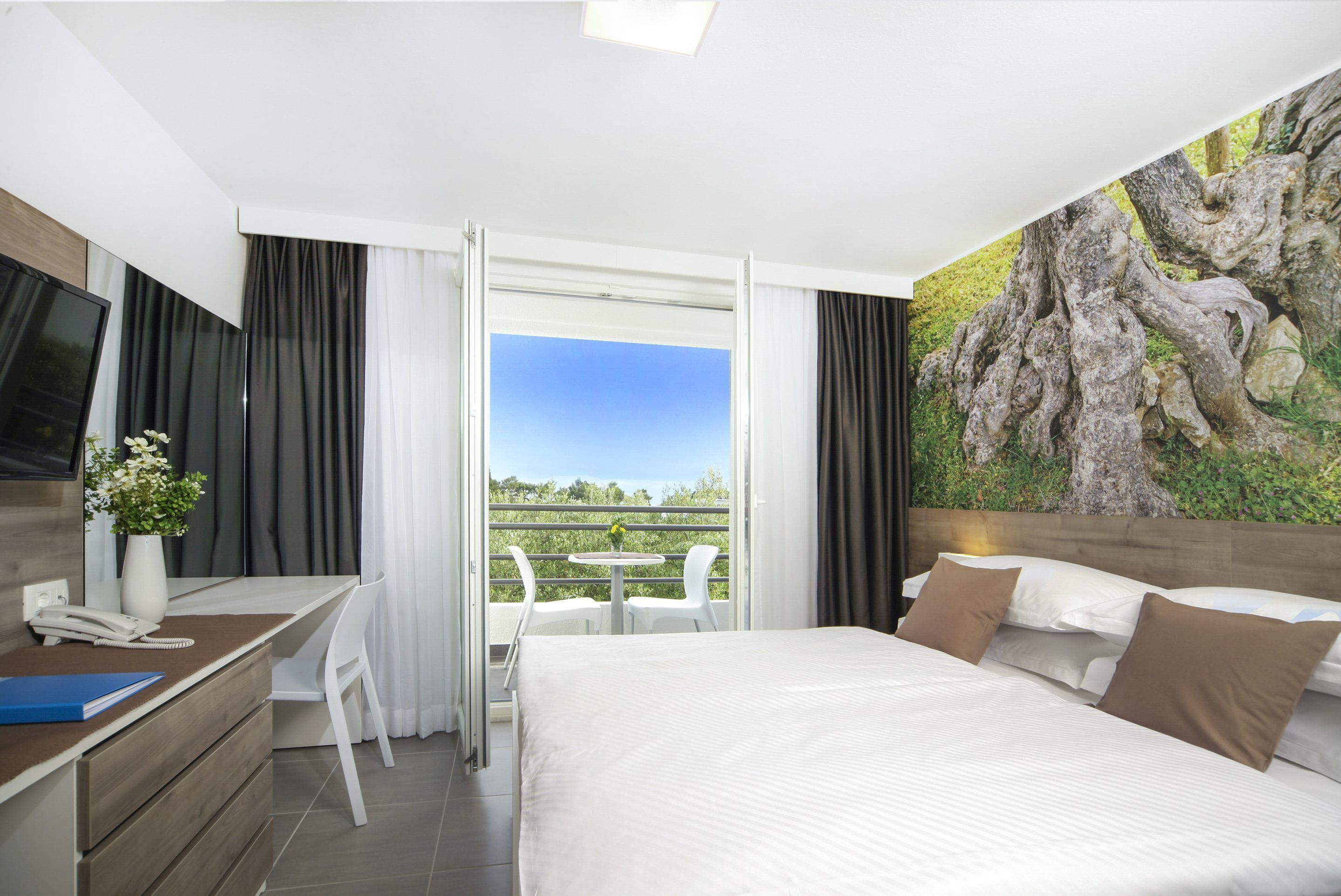 A hotel room with a bed, a desk with a phone, a chair, a small table with chairs, and a large window with a view of greenery.