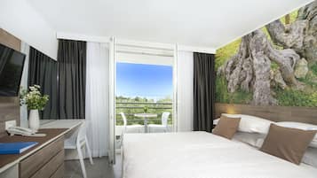 Habitación de hotel con cama, escritorio con teléfono, una silla, una pequeña mesa con sillas y un ventanal con vistas a zonas verdes.