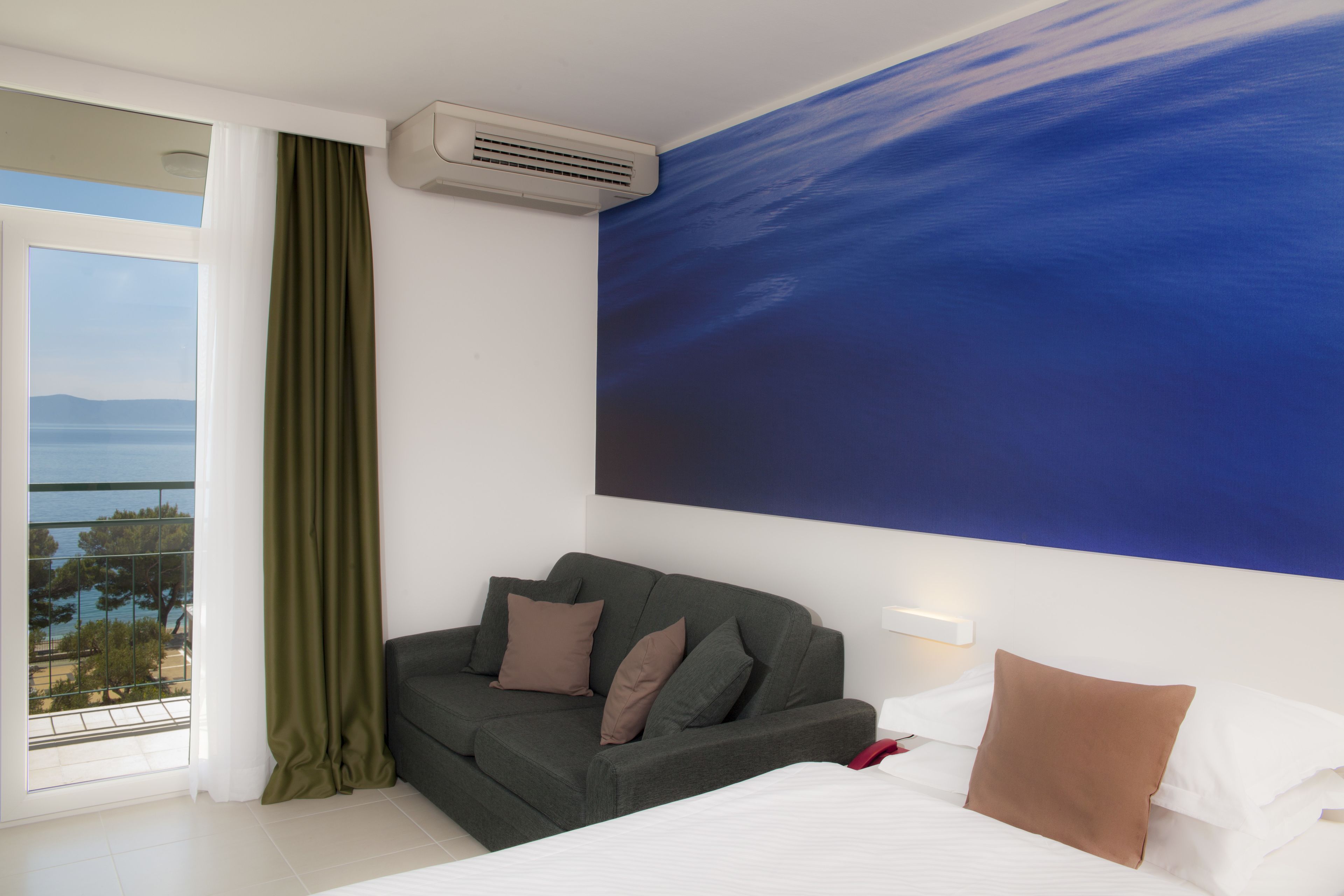 Une chambre d’hôtel équipée d’un lit, d’un canapé, d’une grande fenêtre donnant sur la mer et d’un appareil de climatisation.