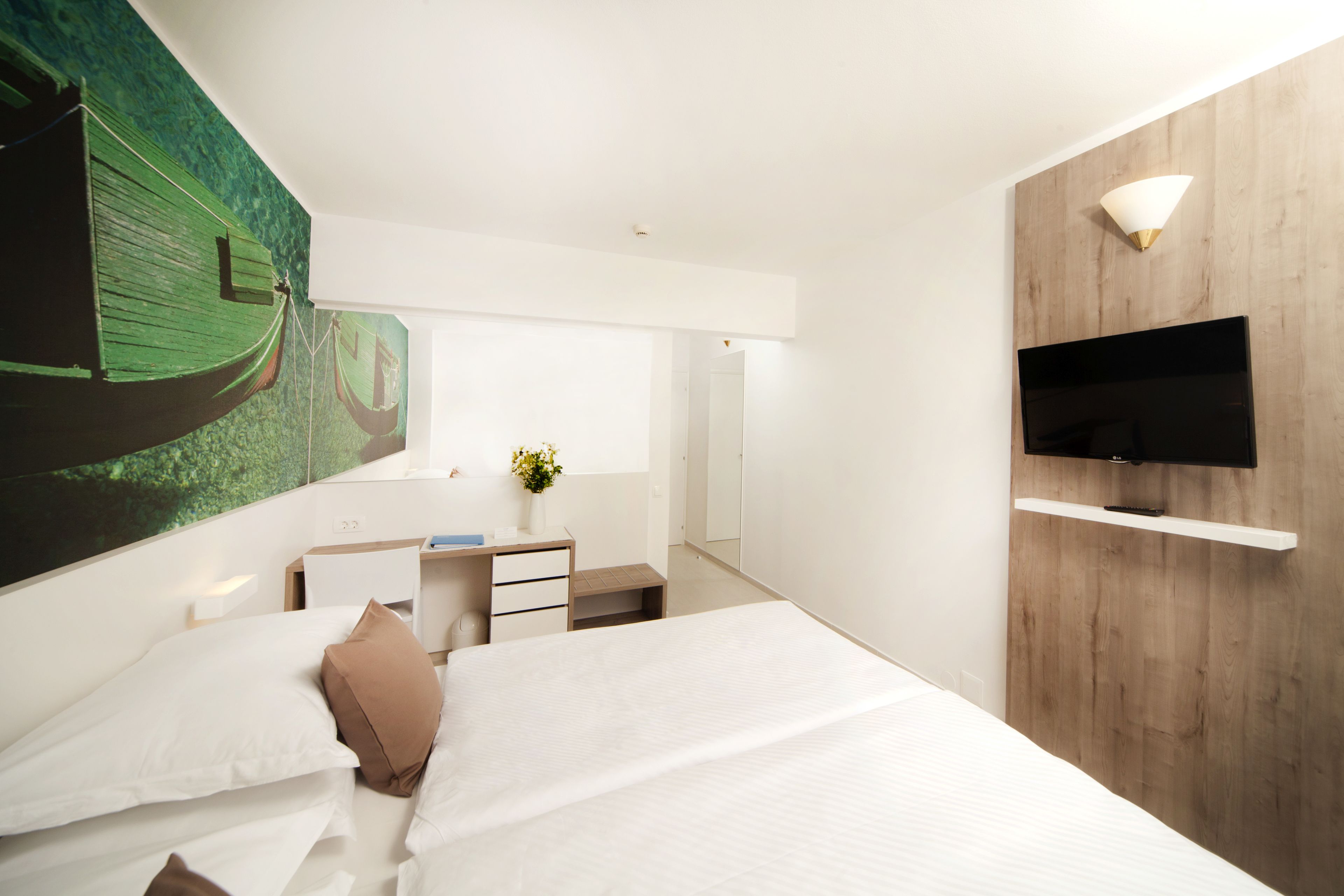 A modern hotel room with a large bed, a desk with a chair, a flat-screen TV mounted on a wooden wall, and a large painting of a boat on the wall.
