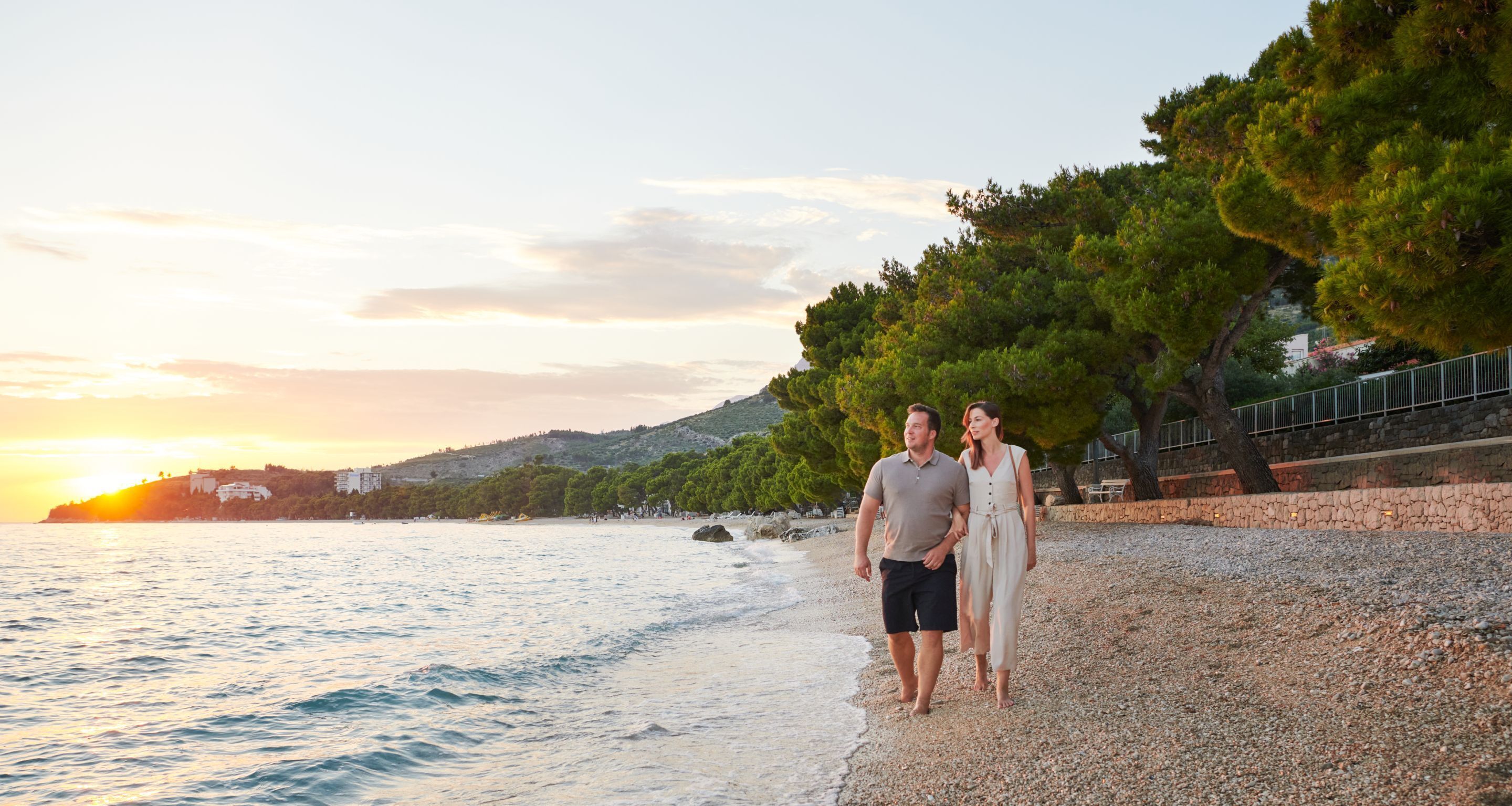 A couple walking on a pebbled beach with a sunset in the background.