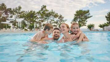 Una piscina con gente nadando, rodeada de árboles y sillas de descanso.