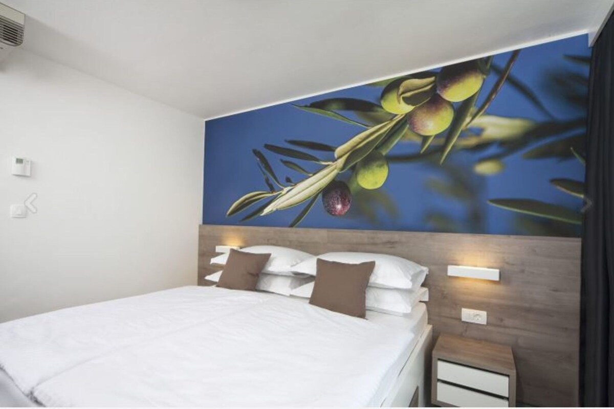 A modern hotel room with a large bed, a wooden headboard, and a wall mural of olives.