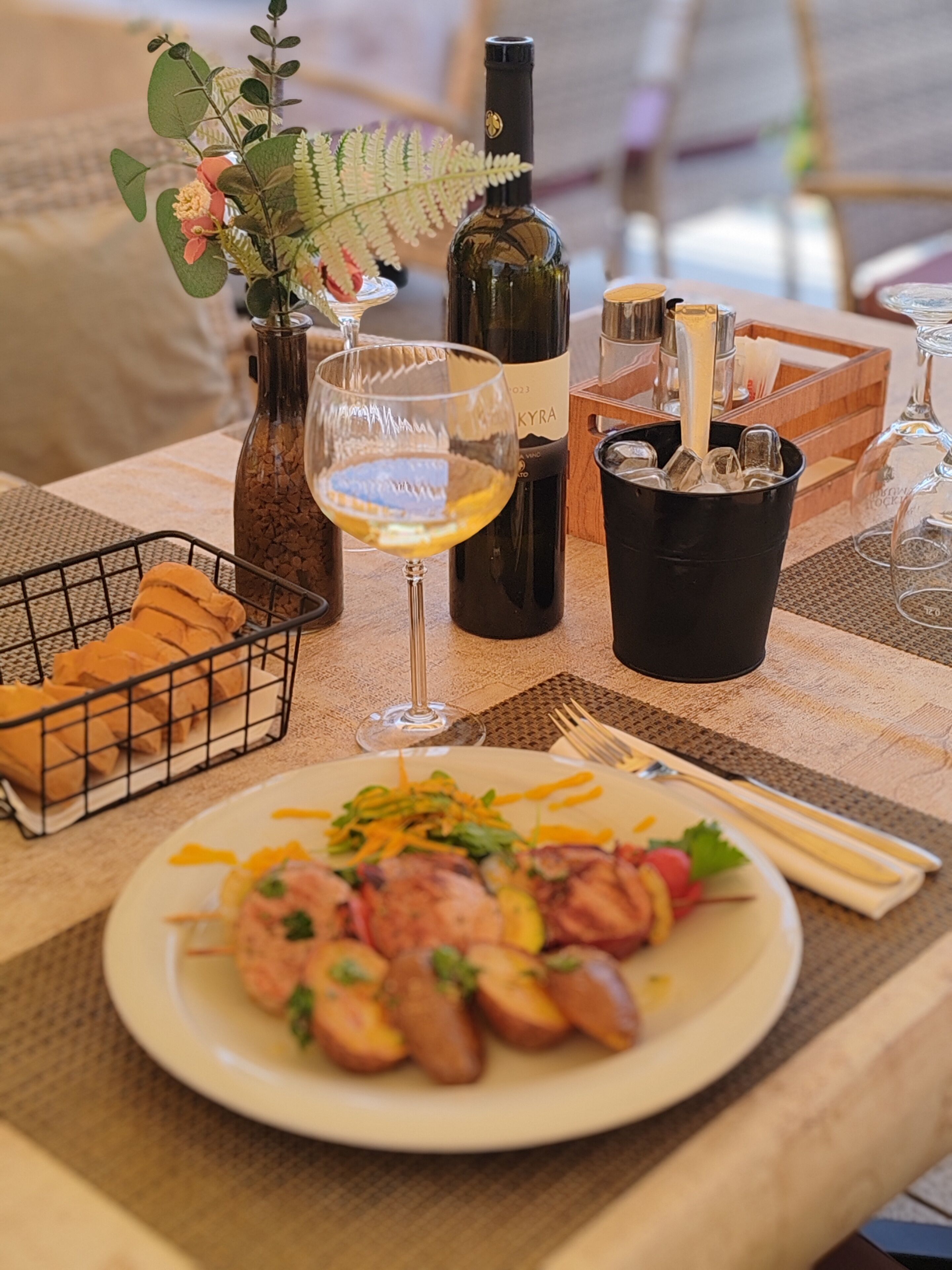 A dining table with a plate of food, a wine bottle, a glass of wine, and a basket of bread.