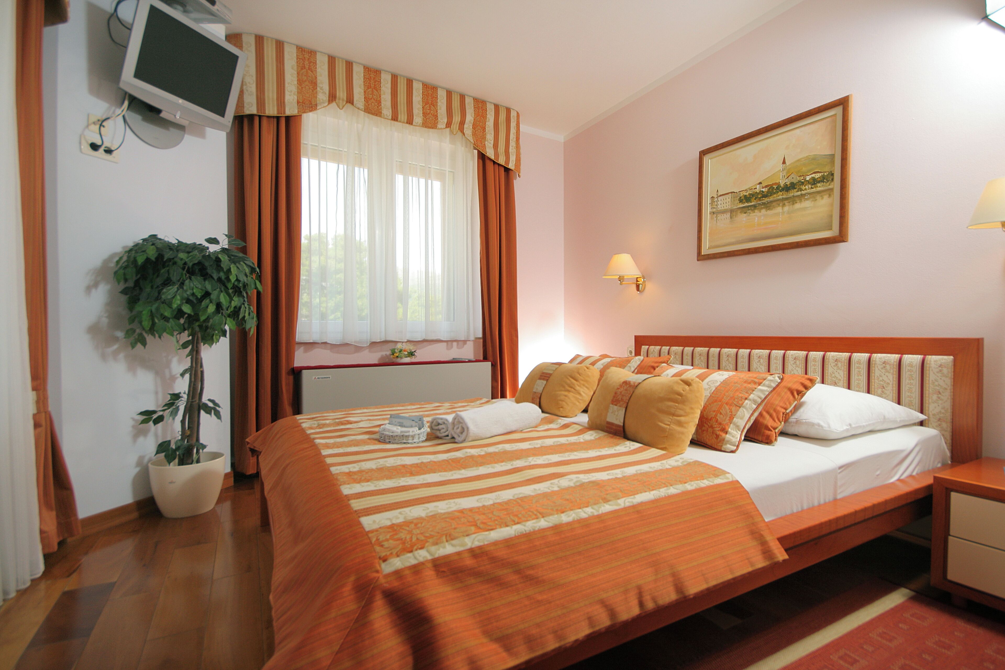 A hotel room with a bed, a potted plant, a painting on the wall, and a television mounted on the wall.