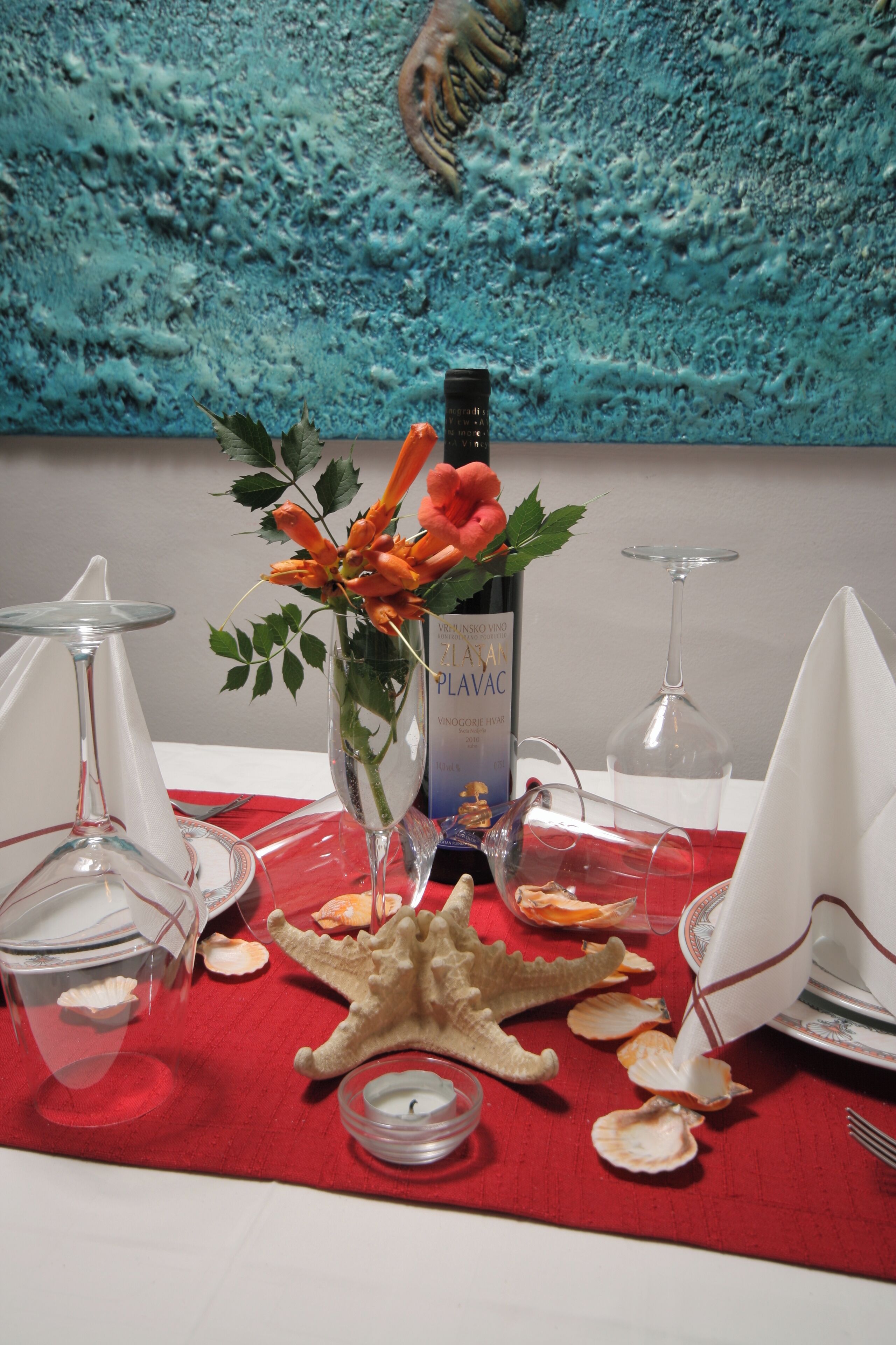 Una mesa dispuesta con una botella de vino, un florero con flores, conchas marinas y una estrella de mar sobre un mantel rojo.
