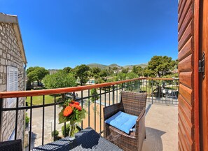 Ein Balkon mit Korbmöbeln, einer Blumenvase und Blick auf eine grüne Landschaft mit Bäumen und Hügeln.