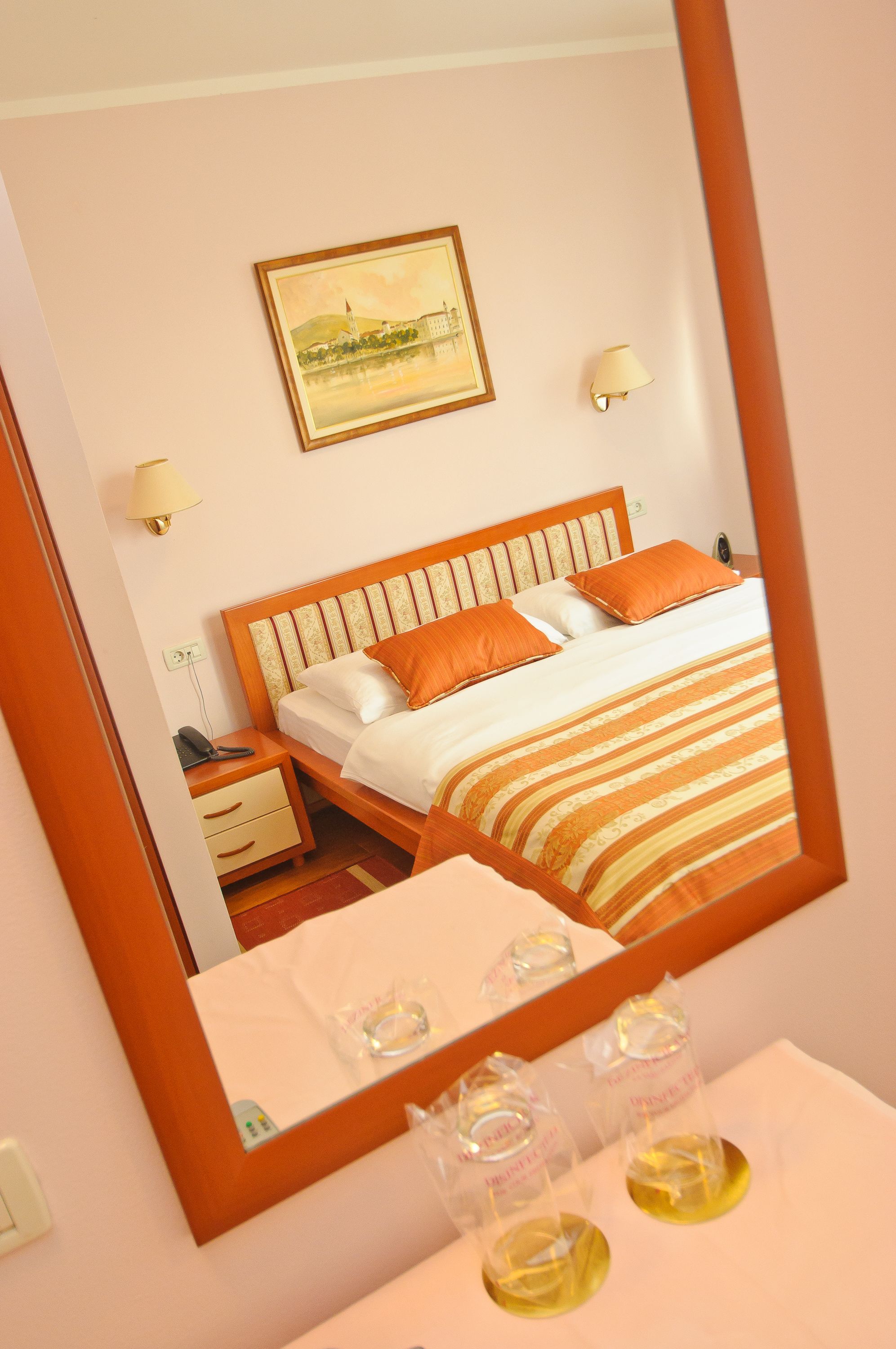 A hotel room with a wooden bed, white bedding, and a painting on the wall. There are two bedside lamps and a nightstand with a phone and a lamp.