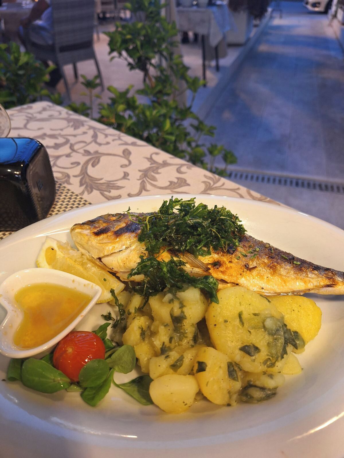 A plate of grilled fish, potatoes, and a small salad with a lemon wedge and a sauce container.