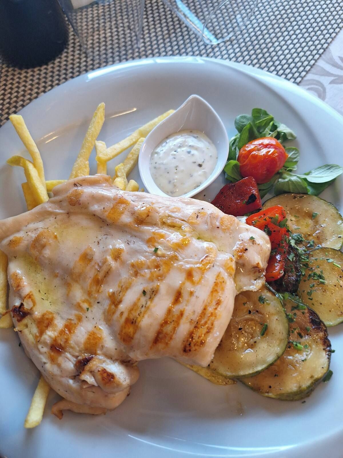 Een bord met gegrilde kip, frieten, een salade en een klein schaaltje saus.