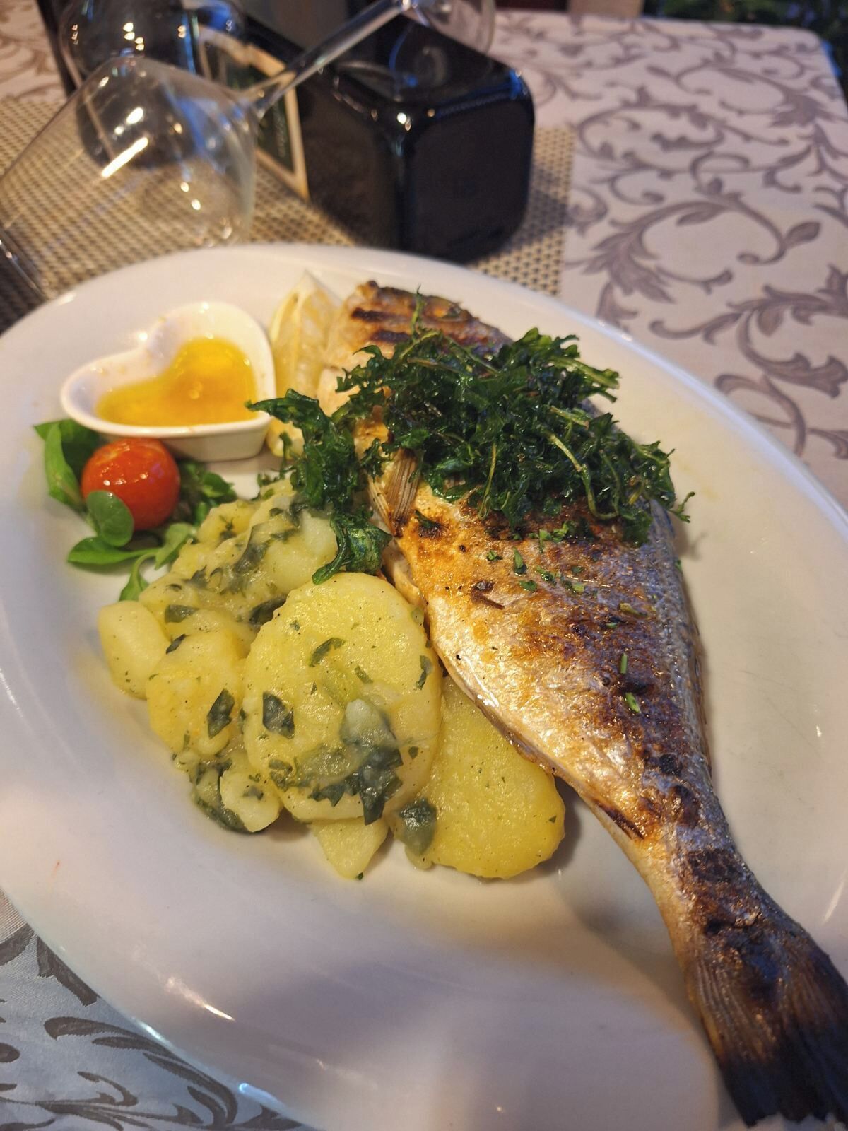 Un plato con pescado a la plancha, papas hervidas, un tomate cherry y un recipiente pequeño con salsa.