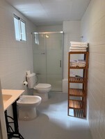 Egy kis fürdőszoba, amelyben WC, bidé és üvegfalas zuhanyzó található. Egy fa polcon törölközők és fürdőszertartozékok vannak.