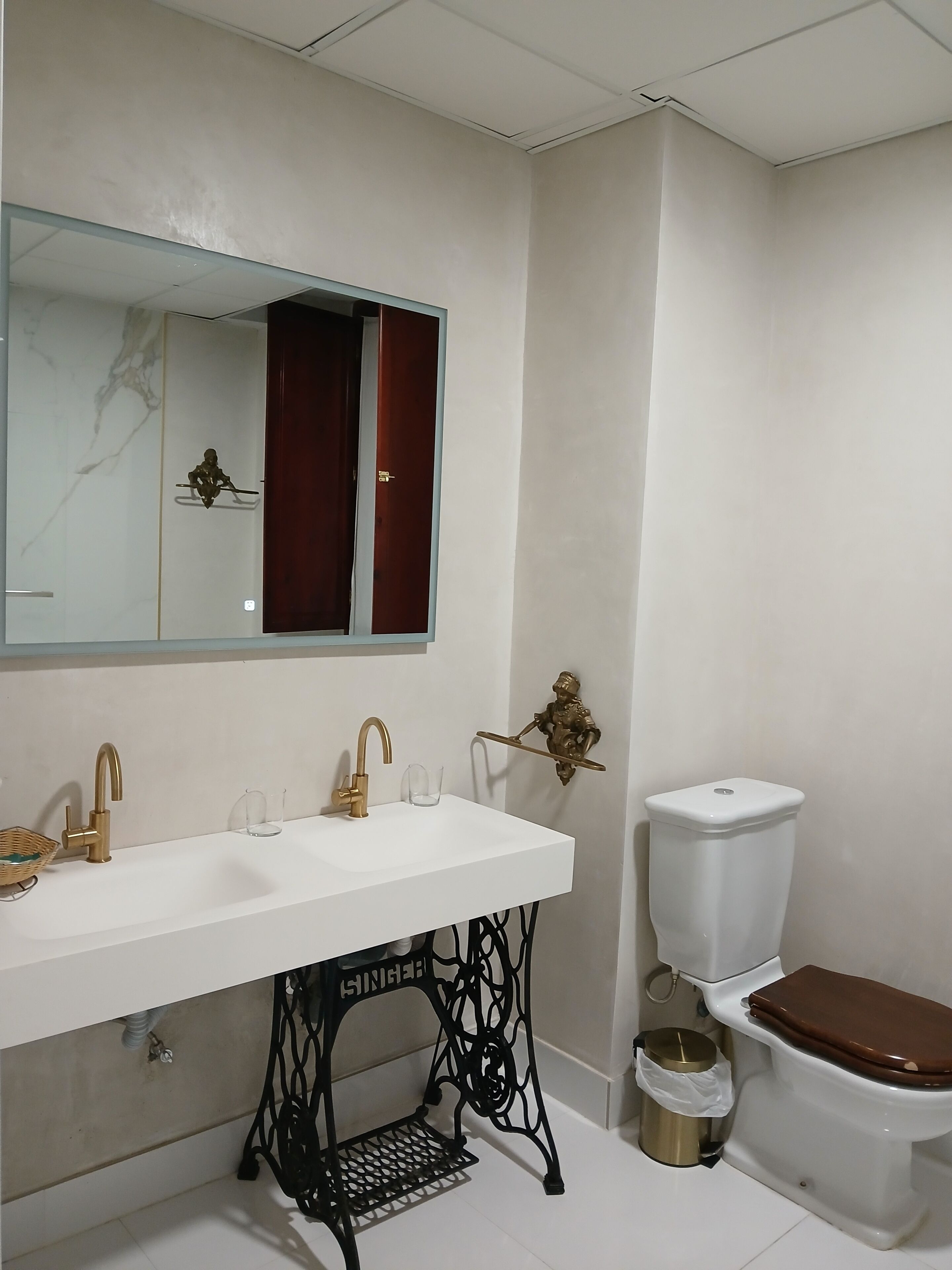 Un baño con lavamanos blanco, grifos dorados, un pedestal metálico negro, inodoro y espejo con cortina roja.