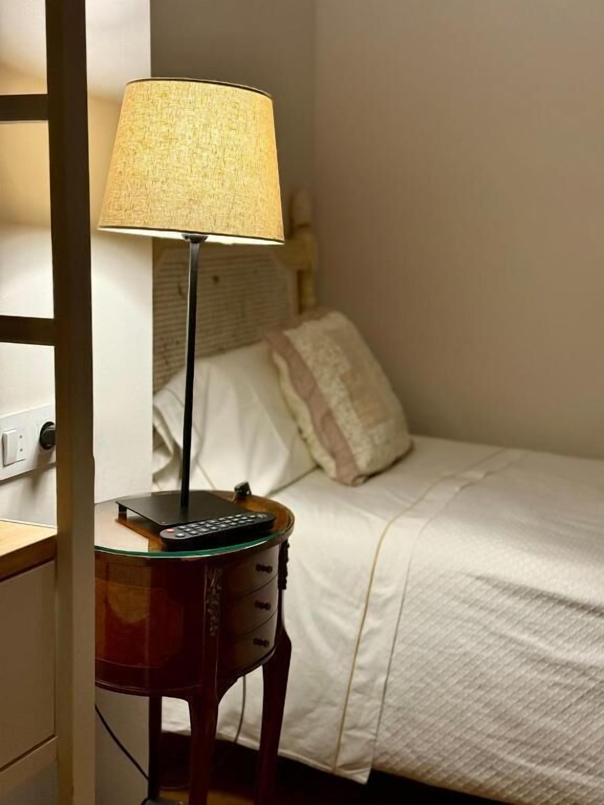 A neatly made bed with a white comforter and a single pillow. A small bedside table with a lamp and a remote control is positioned next to the bed.