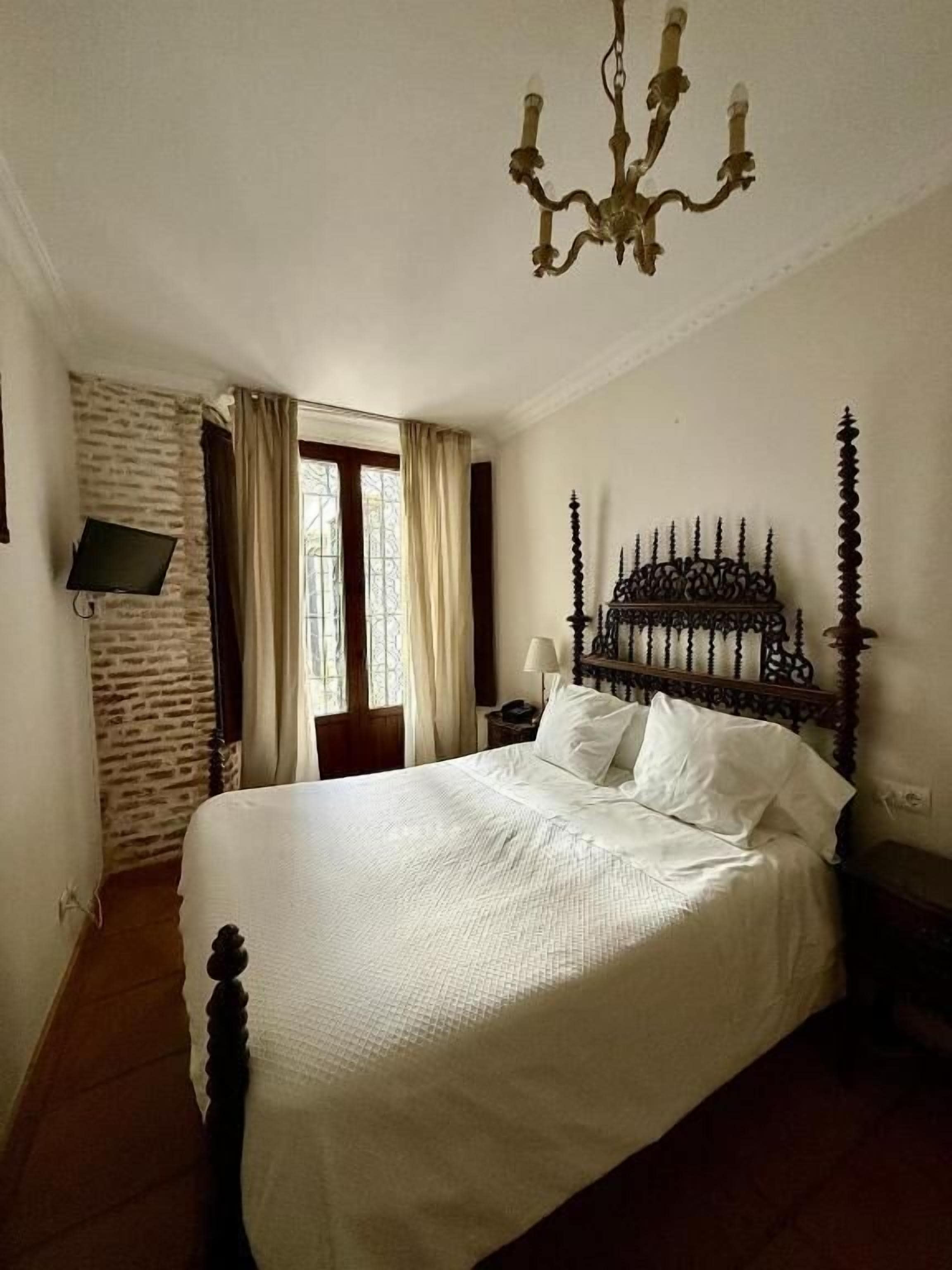 A bedroom with a large bed, a chandelier, a TV mounted on a brick wall, and a window with curtains.