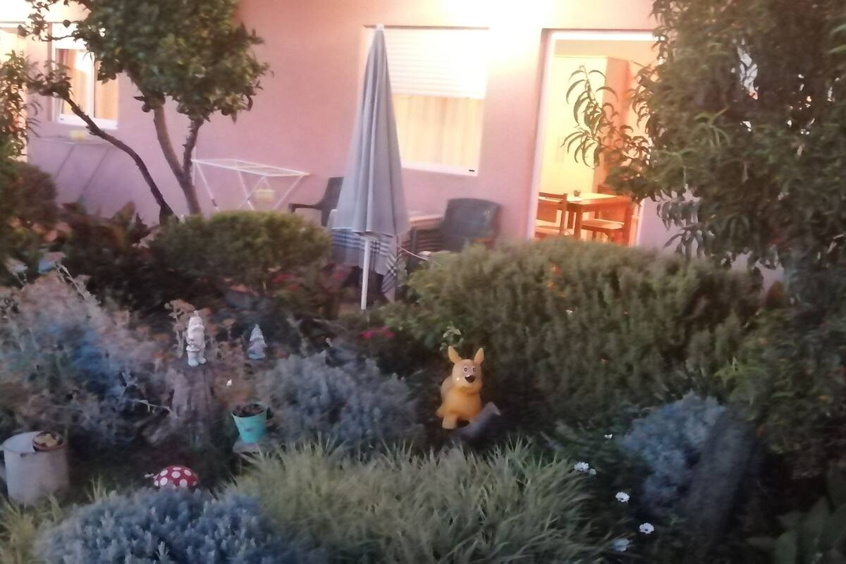 A garden with various plants and a small statue. There is a white chair and a table in the background.