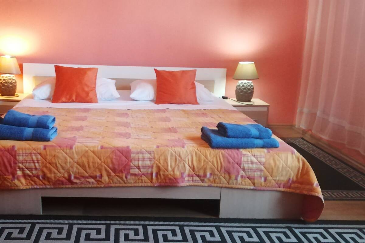 A hotel room with a bed, two nightstands, two lamps, a vase with dried plants, and a patterned rug.