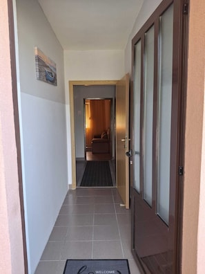 A hallway leading to a room with a bed and a window, a door with a glass panel, and a welcome mat.