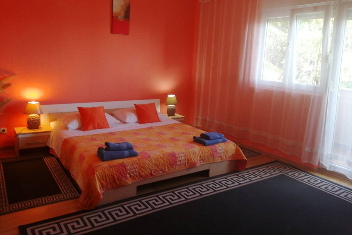 A bedroom with a bed, two bedside lamps, a window with sheer curtains, and a geometric-patterned rug.