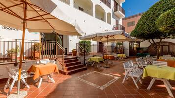 Ein Außenbereich mit einer Terrasse, mehreren Tischen und Stühlen, von denen einige von Sonnenschirmen beschattet werden, und einer Treppe zu einer erhöhten Ebene.