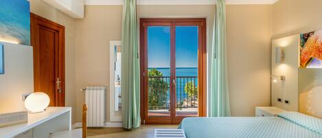 A bedroom with a bed, a desk, a chair, a radiator, and a view of the sea through a door.