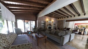 A spacious living room with a stone fireplace, wooden ceiling beams, and a view of the outside. - Troia Tusan Hotel (Çanakkale)