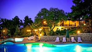 Une piscine avec des chaises longues et un mur de pierre, entourée d’arbres et d’un bâtiment dont les fenêtres sont illuminées.