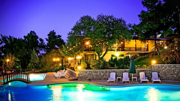 A swimming pool with lounge chairs and a stone wall, surrounded by trees and a building with illuminated windows. - Troia Tusan Hotel (Çanakkale)